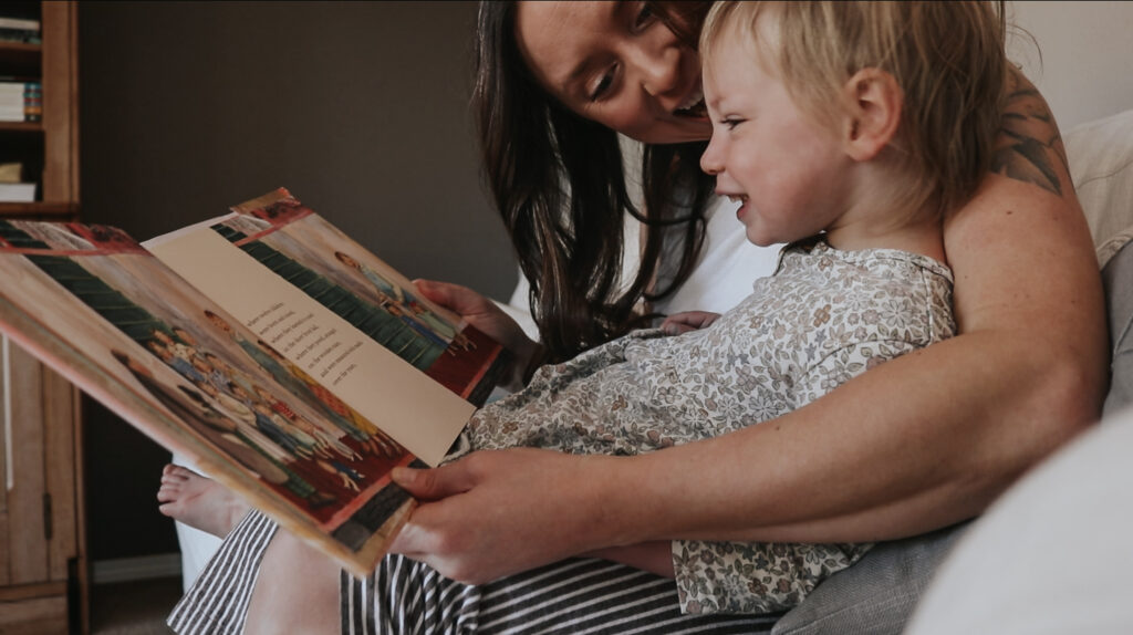 Christtian homemaker holds her toddler daughter in her lap smiling at her while reading a book